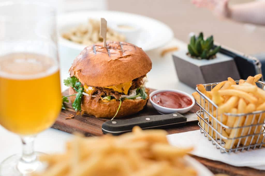 A juicy looking hamburger on a wooden tray next to a basket of fries and a pint of beer.