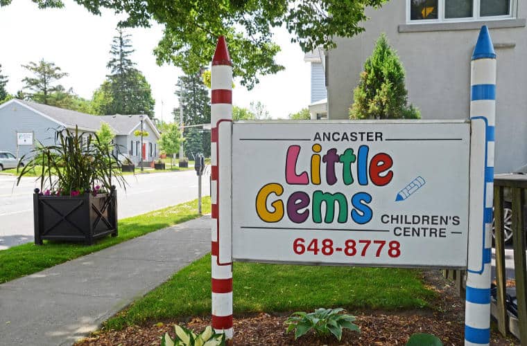 the sign for Ancaster Little Gens on a lawn by the sidewalk.