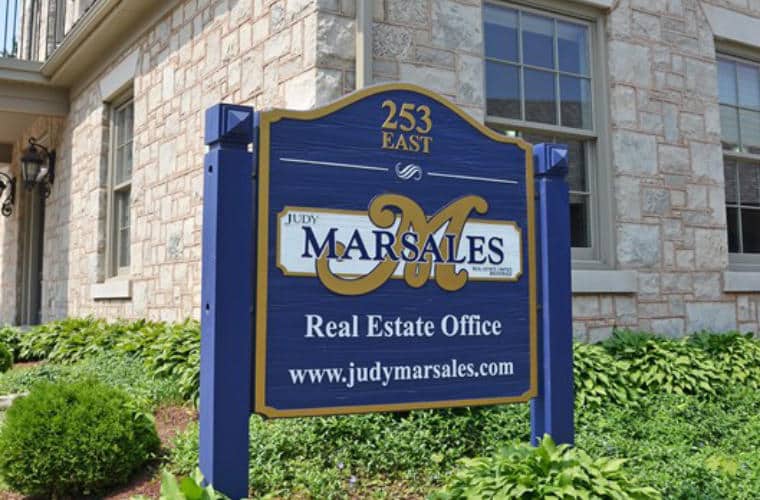 A rough hewn beige stone building with sign for Judy Marsales Real Estate Ltd. on the front lawn.