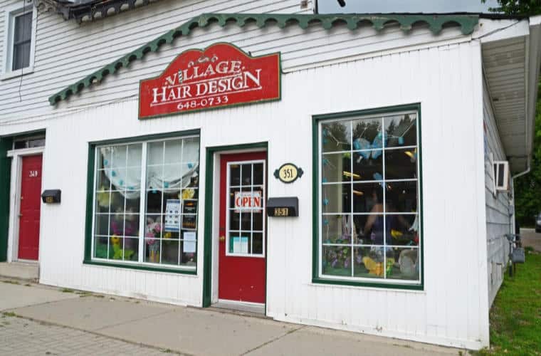 A white building with a red door and large windows with a sign for Village Hair Design above the front entrance.